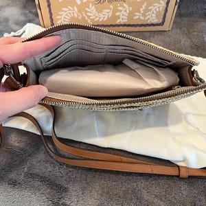 May include: Open view of a beige and brown crossbody bag with a long brown strap. The bag has a zipper closure and a patterned interior with card slots. A box with the Brighton logo is in the background.
