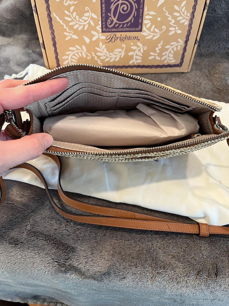 May include: Open view of a beige and brown crossbody bag with a long brown strap. The bag has a zipper closure and a patterned interior with card slots. A box with the Brighton logo is in the background.