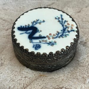 May include: A round, decorative trinket box with a white porcelain lid featuring a blue and floral design. The box has a silver-colored metal base with ornate detailing and a scalloped edge. The lid has a dark border.