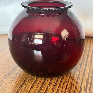 May include: A round, ruby red glass vase with a scalloped rim. The vase has a spherical shape and a glossy finish, reflecting light. The base of the vase has a subtle swirl pattern. This decorative vase is suitable for home decor.