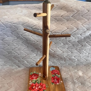 May include: A wooden mug tree with a central post and multiple extending branches for hanging mugs. The square base features a painted floral design with red and blue flowers. A clear, ribbed glass finial sits atop the central wooden post.