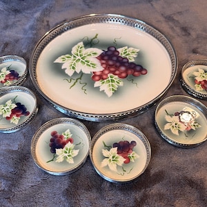 May include: A vintage serving tray and matching coasters set. The tray and coasters feature a painted design of purple grapes and green leaves. The tray has a silver metal rim, and the coasters have a similar silver metal edge.