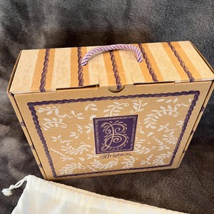 May include: A rectangular cardboard box with a purple rope handle. The box has a beige background with vertical stripes and a decorative border. The front of the box features a purple square with the Brighton logo and the word "Brighton". A white drawstring bag is in front of the box.
