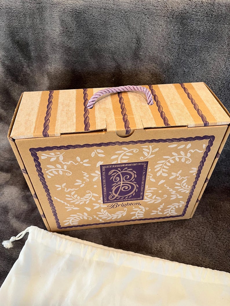 May include: A rectangular cardboard box with a purple rope handle. The box has a beige background with vertical stripes and a decorative border. The front of the box features a purple square with the Brighton logo and the word "Brighton". A white drawstring bag is in front of the box.