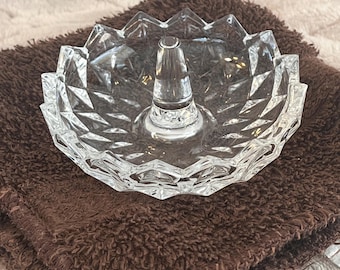 May include: A clear glass ring holder dish featuring a faceted, diamond-cut pattern along the outer rim and a central conical post for storing rings. The dish has a scalloped edge and a transparent, reflective surface throughout the design.