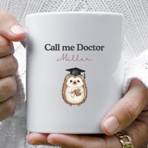 Könnte beinhalten: Weiße Keramik-Tasse mit dem Text "Call me Doctor Müller" in schwarzer und brauner Schrift. Auf der Tasse ist ein Cartoon-Igel mit Doktorhut und Diplom abgebildet.