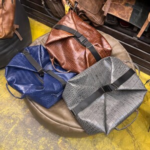 K&ouml;nnte beinhalten: Drei strukturierte Taschen in Braun, Blau und Grau, jeweils mit einer strukturierten Krokodilpr&auml;gung. Jede Tasche ist mit einem schwarzen Nylongurt und einer seitlichen Steckschlie&szlig;e gesichert und verf&uuml;gt &uuml;ber seitliche Tragegriffe.