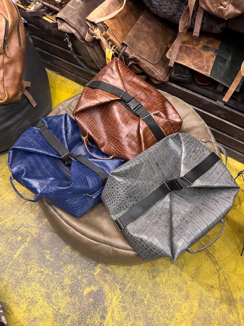 K&ouml;nnte beinhalten: Drei strukturierte Taschen in Braun, Blau und Grau, jeweils mit einer strukturierten Krokodilpr&auml;gung. Jede Tasche ist mit einem schwarzen Nylongurt und einer seitlichen Steckschlie&szlig;e gesichert und verf&uuml;gt &uuml;ber seitliche Tragegriffe.