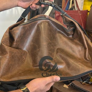 K&ouml;nnte beinhalten: Eine braune Ledertasche mit Used-Optik, ausgestattet mit einem schwarzen Rei&szlig;verschluss, einem schwarzen verstellbaren Riemen mit Kunststoffschnalle und einem schwarzen Logoaufdruck auf der Vorderseite, der "Cheeks" unter einem stilisierten Symbol zeigt.
