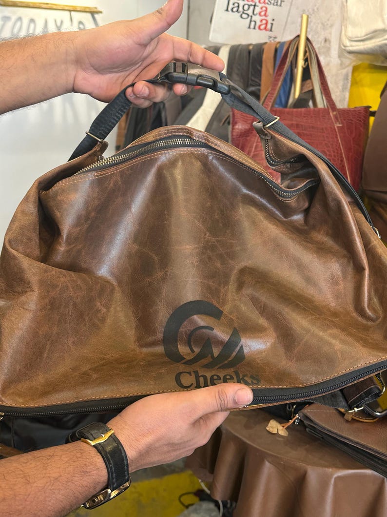 K&ouml;nnte beinhalten: Eine braune Ledertasche mit Used-Optik, ausgestattet mit einem schwarzen Rei&szlig;verschluss, einem schwarzen verstellbaren Riemen mit Kunststoffschnalle und einem schwarzen Logoaufdruck auf der Vorderseite, der "Cheeks" unter einem stilisierten Symbol zeigt.