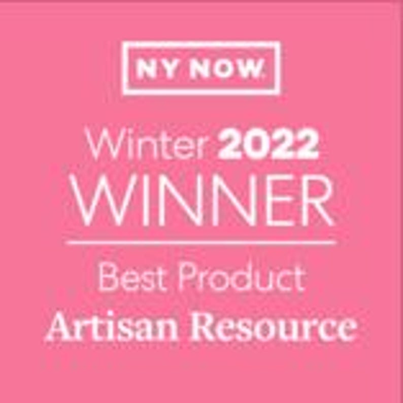 K&ouml;nnte beinhalten: Ein quadratisches, rosafarbenes Grafikschild mit wei&szlig;em Text, der NY NOW, Winter 2022 WINNER, Best Product und Artisan Resource lautet. Das Design ist zentriert mit einem wei&szlig;en rechteckigen Rahmen um den oberen Text und einer Linie unter WINNER.
