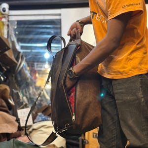 K&ouml;nnte beinhalten: Ein brauner Lederrucksack mit Tragegriff, verstellbaren Schultergurten und einem seitlichen Rei&szlig;verschluss, der ein leuchtend rotes, gemustertes Innenfutter freigibt. Die Tasche hat eine glatte, matte Oberfl&auml;che und metallische Beschl&auml;ge.