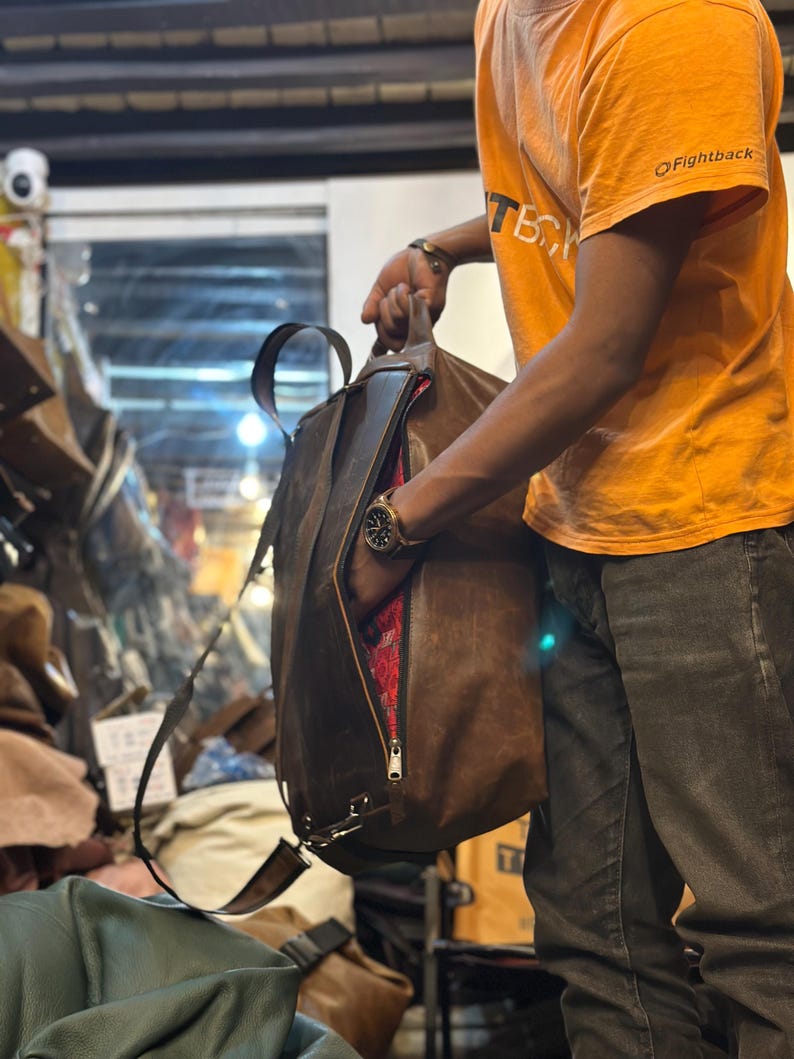 K&ouml;nnte beinhalten: Ein brauner Lederrucksack mit Tragegriff, verstellbaren Schultergurten und einem seitlichen Rei&szlig;verschluss, der ein leuchtend rotes, gemustertes Innenfutter freigibt. Die Tasche hat eine glatte, matte Oberfl&auml;che und metallische Beschl&auml;ge.