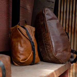 K&ouml;nnte beinhalten: Zwei braune Ledertaschen sind abgebildet. Eine ist ein weicher, formloser Rucksack mit einem vertikalen schwarzen Riemen und Schnallenverschluss. Die andere ist eine gr&ouml;&szlig;ere, strukturierte, dunkelbraune Tasche mit glatter Oberfl&auml;che, seitlichem Rei&szlig;verschluss und schwarzen Riemen.