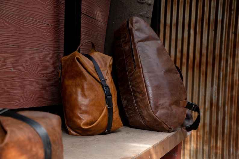 K&ouml;nnte beinhalten: Zwei braune Ledertaschen sind abgebildet. Eine ist ein weicher, formloser Rucksack mit einem vertikalen schwarzen Riemen und Schnallenverschluss. Die andere ist eine gr&ouml;&szlig;ere, strukturierte, dunkelbraune Tasche mit glatter Oberfl&auml;che, seitlichem Rei&szlig;verschluss und schwarzen Riemen.