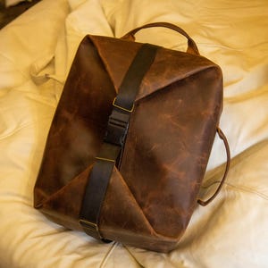 K&ouml;nnte beinhalten: Ein brauner Lederrucksack mit einem vertikalen schwarzen Nylongurt und einem zentralen Kunststoff-Schnallenverschluss. Die Tasche hat eine strukturierte rechteckige Form mit einem Tragegriff und Seitengurten sowie eine abgenutzte Lederoberfl&auml;che.