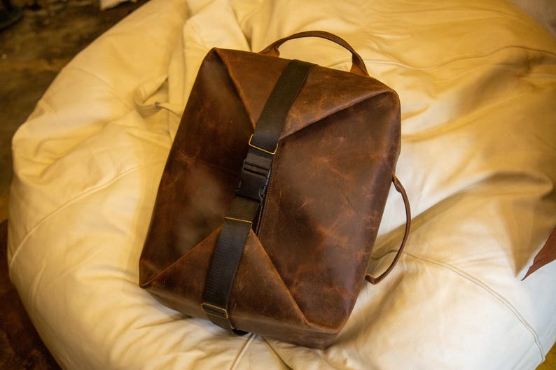 K&ouml;nnte beinhalten: Ein brauner Lederrucksack mit einem vertikalen schwarzen Nylongurt und einem zentralen Kunststoff-Schnallenverschluss. Die Tasche hat eine strukturierte rechteckige Form mit einem Tragegriff und Seitengurten sowie eine abgenutzte Lederoberfl&auml;che.