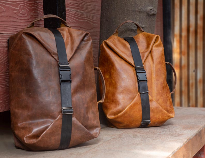 May include: Two brown leather backpacks with a smooth, matte finish. Each bag features a vertical black nylon strap secured with a central plastic buckle, a top handle, and a side handle. The leather has a natural, slightly distressed texture.