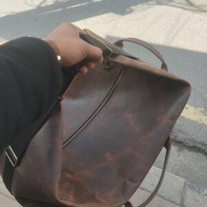 K&ouml;nnte beinhalten: Eine dunkelbraune Reisetasche aus Leder mit einem gro&szlig;en zentralen Rei&szlig;verschlussfach, zwei Tragegriffen an der Oberseite und einem verstellbaren schwarzen Schultergurt mit silberfarbenen Metallbeschl&auml;gen. Die Lederoberfl&auml;che zeigt eine nat&uuml;rliche Textur.