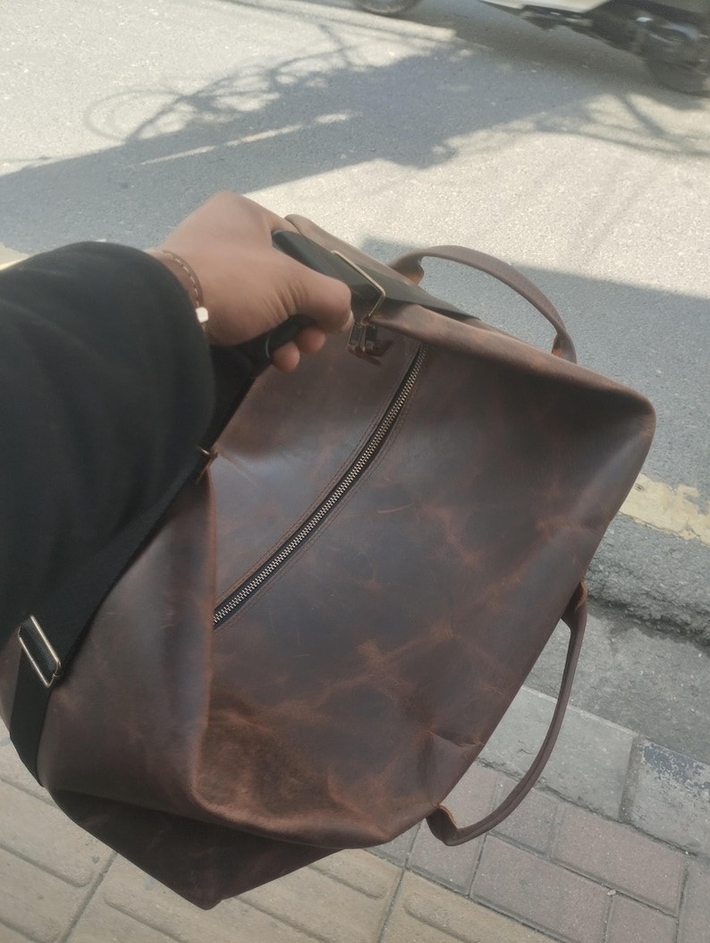 K&ouml;nnte beinhalten: Eine dunkelbraune Reisetasche aus Leder mit einem gro&szlig;en zentralen Rei&szlig;verschlussfach, zwei Tragegriffen an der Oberseite und einem verstellbaren schwarzen Schultergurt mit silberfarbenen Metallbeschl&auml;gen. Die Lederoberfl&auml;che zeigt eine nat&uuml;rliche Textur.