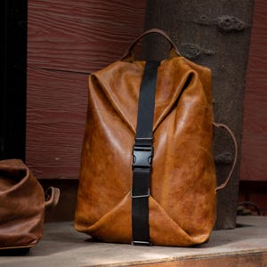 K&ouml;nnte beinhalten: Ein beigefarbener Lederrucksack mit einem vertikalen schwarzen Nylongurt und einem seitlichen Kunststoff-Steckverschluss. Die Tasche verf&uuml;gt &uuml;ber einen Tragegriff oben, einen Seitengriff und eine weiche, unstrukturierte Form mit Falten.