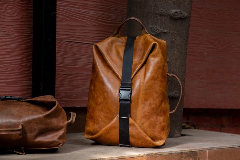 K&ouml;nnte beinhalten: Ein beigefarbener Lederrucksack mit einem vertikalen schwarzen Nylongurt und einem seitlichen Kunststoff-Steckverschluss. Die Tasche verf&uuml;gt &uuml;ber einen Tragegriff oben, einen Seitengriff und eine weiche, unstrukturierte Form mit Falten.