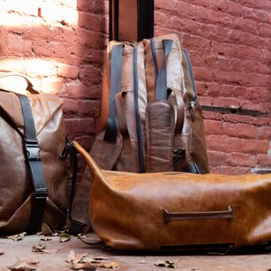 K&ouml;nnte beinhalten: Drei braune Ledertaschen mit glatter Textur und schwarzen Riemen. Ein aufrechter Rucksack hat einen vertikalen schwarzen Schnallenriemen, ein weiterer h&ouml;herer Rucksack steht dahinter und eine horizontale Reisetasche mit Griff liegt im Vordergrund.