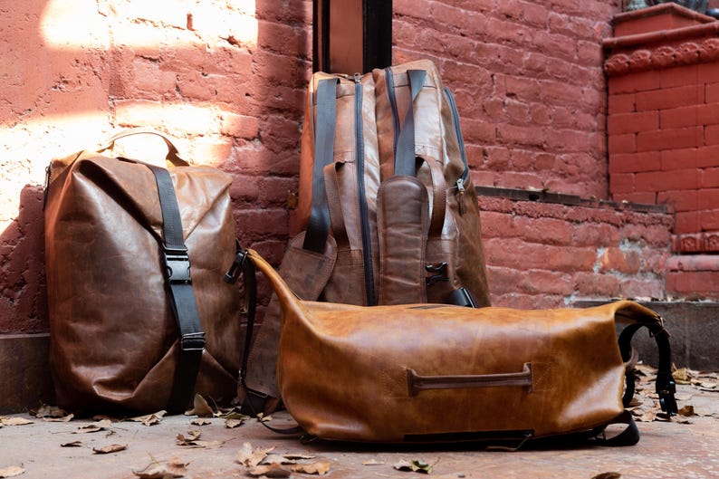K&ouml;nnte beinhalten: Drei braune Ledertaschen mit glatter Textur und schwarzen Riemen. Ein aufrechter Rucksack hat einen vertikalen schwarzen Schnallenriemen, ein weiterer h&ouml;herer Rucksack steht dahinter und eine horizontale Reisetasche mit Griff liegt im Vordergrund.