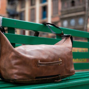 K&ouml;nnte beinhalten: Eine braune Reisetasche aus Leder mit einem horizontalen Griff auf der Vorderseite, einem schwarzen Rei&szlig;verschluss an der Unterseite und verstellbaren schwarzen Schultergurten. Die Tasche hat eine weiche, unstrukturierte Form mit sichtbaren N&auml;hten.