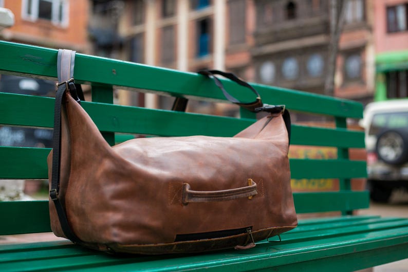 K&ouml;nnte beinhalten: Eine braune Reisetasche aus Leder mit einem horizontalen Griff auf der Vorderseite, einem schwarzen Rei&szlig;verschluss an der Unterseite und verstellbaren schwarzen Schultergurten. Die Tasche hat eine weiche, unstrukturierte Form mit sichtbaren N&auml;hten.