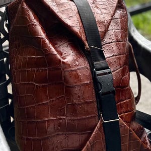 K&ouml;nnte beinhalten: Ein brauner Rucksack mit texturiertem Krokodilledermuster und gl&auml;nzender Oberfl&auml;che. Die Tasche verf&uuml;gt &uuml;ber einen vertikalen schwarzen Nylongurt mit Kunststoff-Steckschnalle und ein kleines goldfarbenes Metall-Logoschild am oberen Griff.