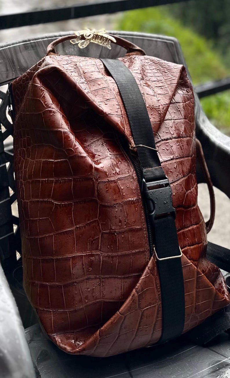 K&ouml;nnte beinhalten: Ein brauner Rucksack mit texturiertem Krokodilledermuster und gl&auml;nzender Oberfl&auml;che. Die Tasche verf&uuml;gt &uuml;ber einen vertikalen schwarzen Nylongurt mit Kunststoff-Steckschnalle und ein kleines goldfarbenes Metall-Logoschild am oberen Griff.
