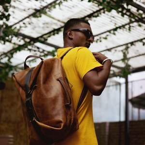 K&ouml;nnte beinhalten: Ein gro&szlig;er Rucksack aus hellbraunem Leder mit einem Tragegriff oben, einem vertikalen dunkelbraunen Riemendetail und einem seitlichen Griff. Die Tasche hat ein strukturiertes, aber flexibles Design mit sichtbaren N&auml;hten und Metallbeschl&auml;gen.
