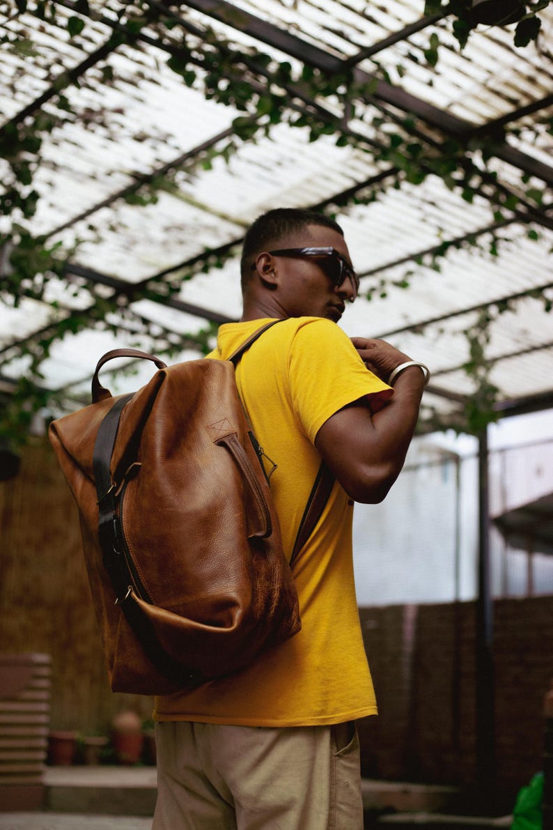 K&ouml;nnte beinhalten: Ein gro&szlig;er Rucksack aus hellbraunem Leder mit einem Tragegriff oben, einem vertikalen dunkelbraunen Riemendetail und einem seitlichen Griff. Die Tasche hat ein strukturiertes, aber flexibles Design mit sichtbaren N&auml;hten und Metallbeschl&auml;gen.