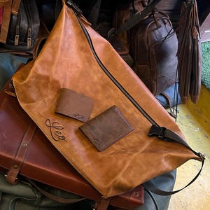 K&ouml;nnte beinhalten: Eine hellbraune Ledertasche mit schwarzem Rei&szlig;verschluss und schwarzer Kunststoffschnalle, auf der vorne der Name Leo eingepr&auml;gt ist. Zwei kleine, dunkelbraune Ledergeldb&ouml;rsen mit den eingepr&auml;gten Initialen LR liegen auf der Tasche.