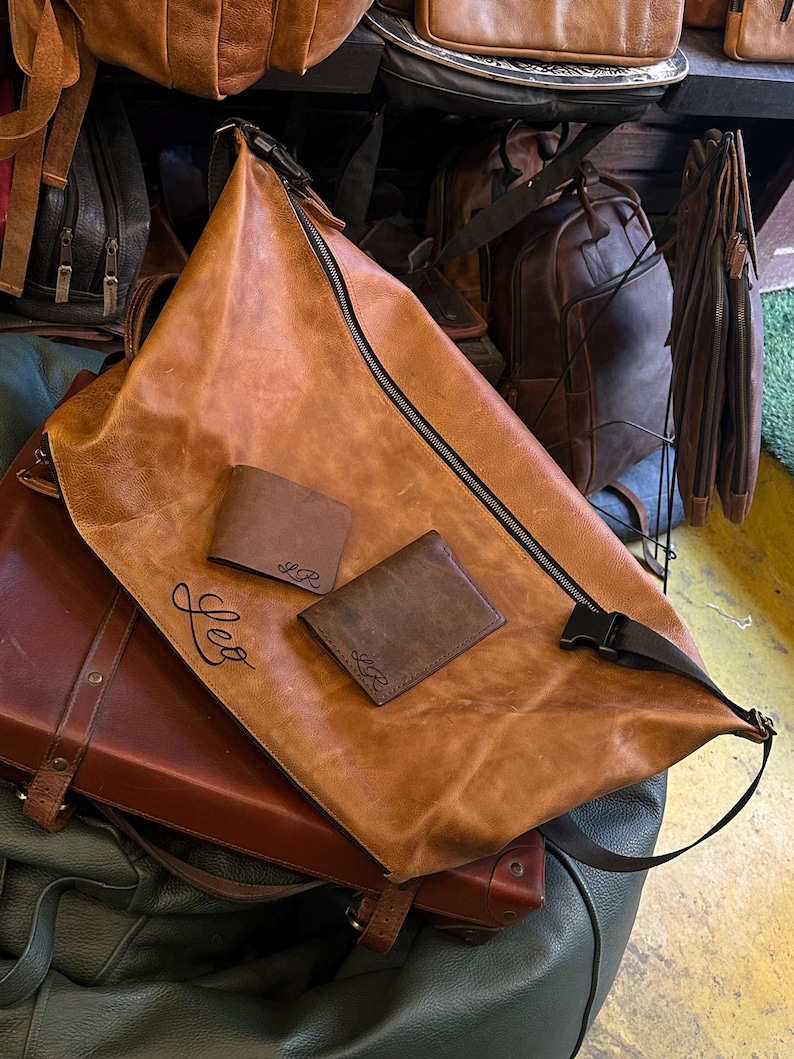 K&ouml;nnte beinhalten: Eine hellbraune Ledertasche mit schwarzem Rei&szlig;verschluss und schwarzer Kunststoffschnalle, auf der vorne der Name Leo eingepr&auml;gt ist. Zwei kleine, dunkelbraune Ledergeldb&ouml;rsen mit den eingepr&auml;gten Initialen LR liegen auf der Tasche.