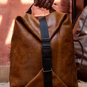 K&ouml;nnte beinhalten: Ein beigefarbener Lederrucksack mit einem vertikalen schwarzen Nylongurt und einem Kunststoff-Steckverschluss. Die Tasche hat eine weiche, unstrukturierte Form mit einem Tragegriff oben und an der Seite sowie eine nat&uuml;rliche Narbung und Farbnuancen.