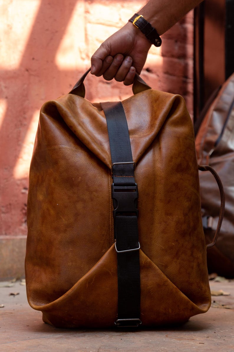 K&ouml;nnte beinhalten: Ein beigefarbener Lederrucksack mit einem vertikalen schwarzen Nylongurt und einem Kunststoff-Steckverschluss. Die Tasche hat eine weiche, unstrukturierte Form mit einem Tragegriff oben und an der Seite sowie eine nat&uuml;rliche Narbung und Farbnuancen.