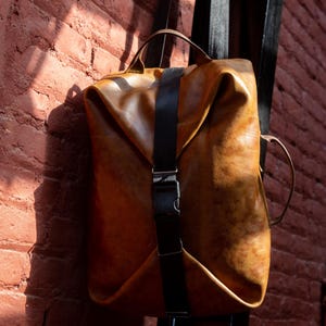 K&ouml;nnte beinhalten: Ein beigefarbener Lederrucksack mit strukturiertem, gefaltetem Design, einem zentralen schwarzen vertikalen Riemen und einer seitlichen Kunststoffschnalle. Die Tasche verf&uuml;gt &uuml;ber einen Tragegriff oben und einen seitlichen Griff mit glatter Oberfl&auml;che.