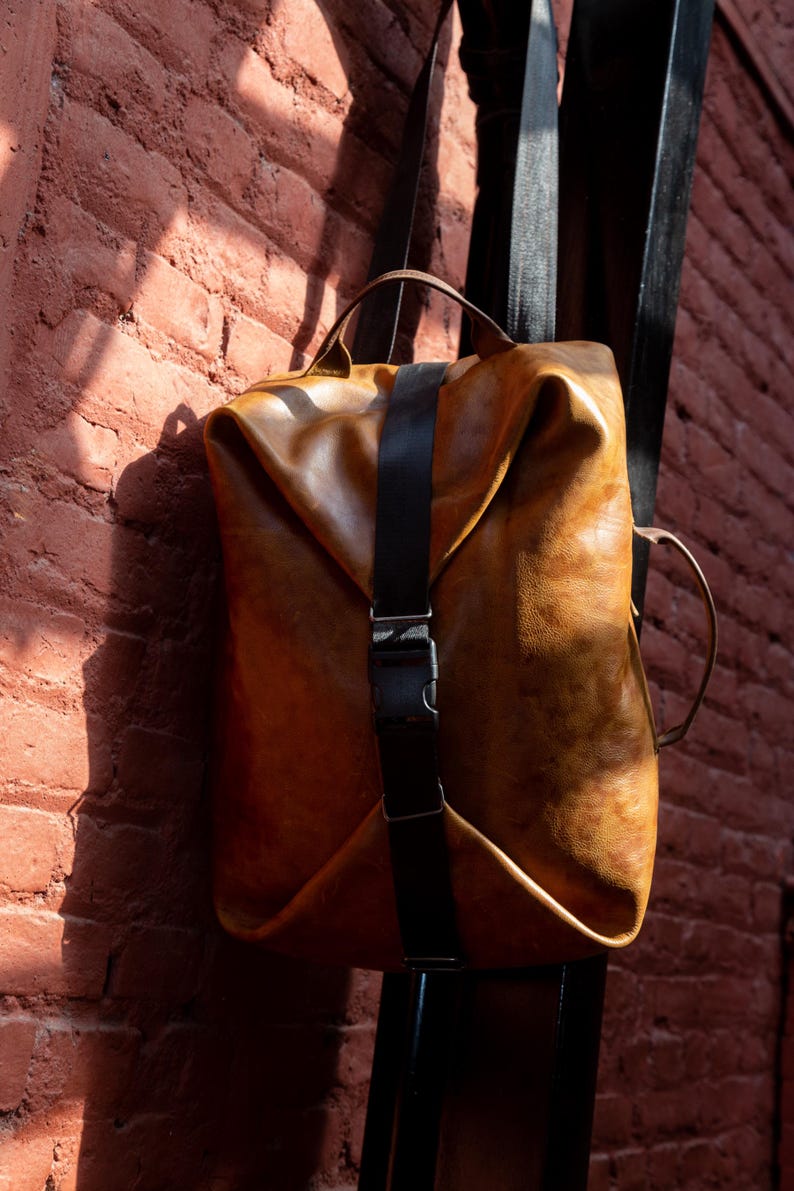 K&ouml;nnte beinhalten: Ein beigefarbener Lederrucksack mit strukturiertem, gefaltetem Design, einem zentralen schwarzen vertikalen Riemen und einer seitlichen Kunststoffschnalle. Die Tasche verf&uuml;gt &uuml;ber einen Tragegriff oben und einen seitlichen Griff mit glatter Oberfl&auml;che.