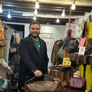 K&ouml;nnte beinhalten: Eine braune Reisetasche aus Leder mit dem eingepr&auml;gten Wort MULCH auf der Vorderseite, gehalten in einem Gesch&auml;ft voller verschiedener Ledertaschen, Rucks&auml;cke und Accessoires, die auf Tischen und Regalen unter h&auml;ngenden Gl&uuml;hbirnen pr&auml;sentiert werden.
