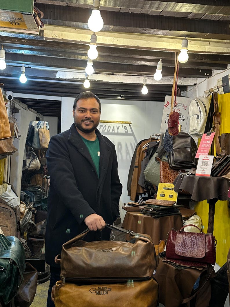 K&ouml;nnte beinhalten: Eine braune Reisetasche aus Leder mit dem eingepr&auml;gten Wort MULCH auf der Vorderseite, gehalten in einem Gesch&auml;ft voller verschiedener Ledertaschen, Rucks&auml;cke und Accessoires, die auf Tischen und Regalen unter h&auml;ngenden Gl&uuml;hbirnen pr&auml;sentiert werden.