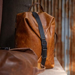K&ouml;nnte beinhalten: Ein beigefarbener Lederrucksack mit einem vertikalen schwarzen Nylongurt und einer Kunststoff-Steckschnalle. Die Tasche hat einen Tragegriff an der Oberseite und eine weiche, unstrukturierte Form. Eine zweite, dunkelbraune Ledertasche ist im Vordergrund zu sehen.