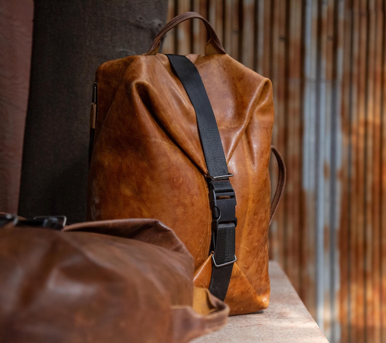 K&ouml;nnte beinhalten: Ein beigefarbener Lederrucksack mit einem vertikalen schwarzen Nylongurt und einer Kunststoff-Steckschnalle. Die Tasche hat einen Tragegriff an der Oberseite und eine weiche, unstrukturierte Form. Eine zweite, dunkelbraune Ledertasche ist im Vordergrund zu sehen.