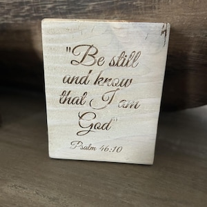 May include: A small, rectangular wooden plaque with the engraved words "Be still and know that I am God" and "Psalm 46:10". The wood has a light, weathered look with visible grain and a slightly distressed finish.