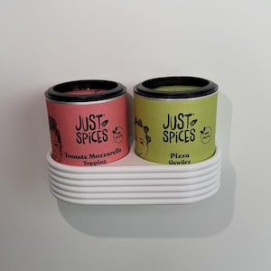 Könnte beinhalten: Zwei Gewürzbehälter in einem weißen Halter. Einer ist rosa mit dem Text "Tomate Mozzarella Topping", der andere grün mit dem Text "Pizza Gewürz". Beide haben schwarze Deckel und den Text "JUST SPICES".