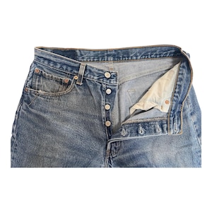 May include: Close-up of blue denim jeans with a button fly. The jeans have a classic five-pocket design, with visible wear and fading. The button fly is partially unbuttoned, revealing the interior lining.