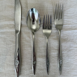 Puede incluir: Un juego de cuatro cubiertos de acero inoxidable que incluye un cuchillo de mesa, una cuchara, un tenedor de ensalada y un tenedor de mesa. Cada pieza tiene un acabado pulido y un motivo de rosa en relieve en la base del mango.