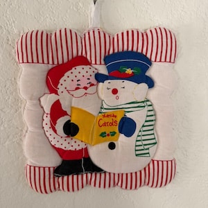 Peut inclure: Une tenture murale carrée en tissu matelassé avec une bordure rayée rouge et blanche. Le centre présente une applique du Père Noël et d'un bonhomme de neige portant un chapeau bleu et une écharpe rayée verte, tenant un livre jaune Yuletide Carols.