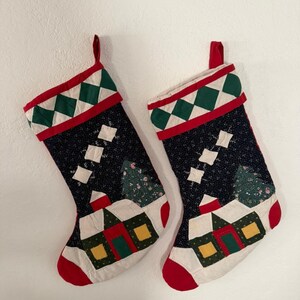 Op de afbeelding: Twee gewatteerde kerstsokken met een donkerblauwe stoffen achtergrond met een kleine bloemenprint. Elke sok toont een huisapplicatie met gele ramen en een rood dak, een groene boom en een boord met een groen-wit ruitpatroon.